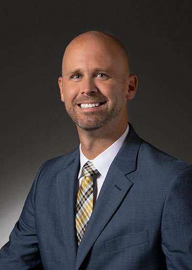 Professional portrait of a man with a shaved head, wearing a suit and tie, smiling against a dark background.