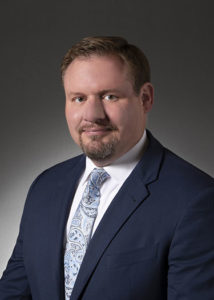 Professional headshot of a man in a dark suit and patterned tie, smiling against a gray background.