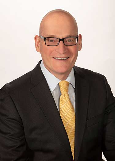 Professional portrait of a man wearing glasses and a suit with a yellow tie, smiling against a light background.