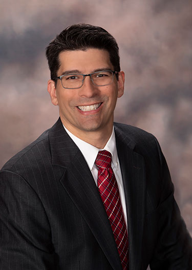 Headshot of a professional man smiling, wearing glasses, a dark suit, and a red tie, with a neutral background.